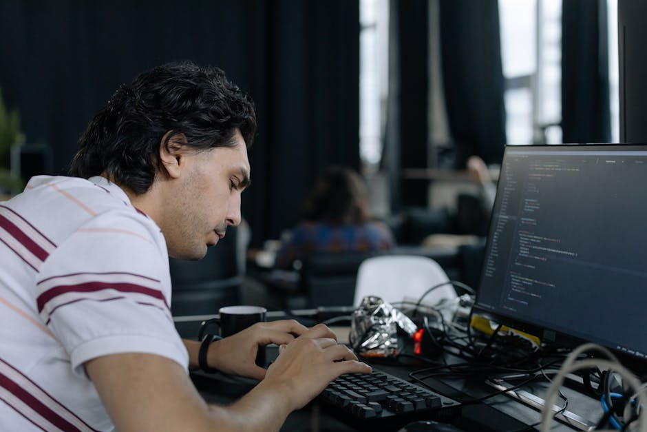A young man with dark, curly hair is seated at a desk in an indoor office environment, focused on his work on a computer with a large monitor displaying lines of code. The workspace features a clutter