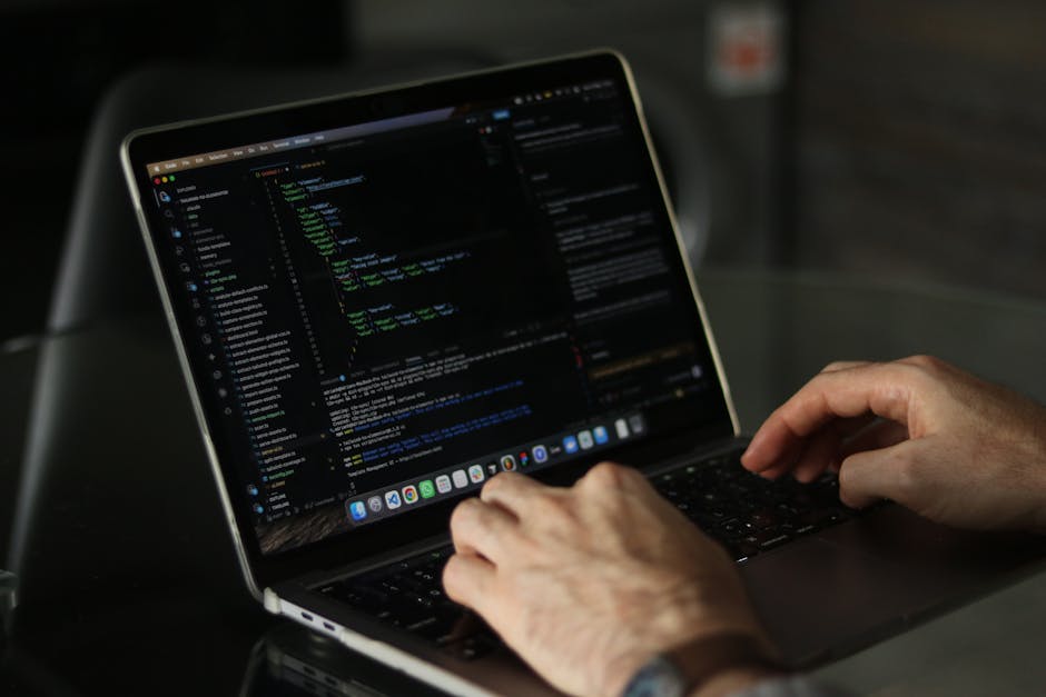 A young man with dark, curly hair is seated at a desk in an indoor office environment, focused on his work on a computer with a large monitor displaying lines of code. The workspace features a clutter